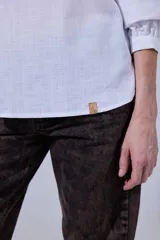 Camisa blanca de algodón con pechera decorada con alforzas y puntillas. Presenta cierre frontal con botones, mangas tres cuartos con frunces y detalles de puntilla, y un diseño de corte holgado con ruedo redondeado.