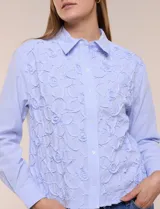 Camisa de algodón celeste con patrón de milrayas finas y aplicaciones florales en relieve deshilachado en el frente. Cuenta con cuello camisero tradicional, cierre frontal con botones, mangas largas con puños abotonados y corte semi holgado.
