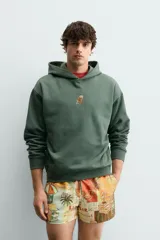 Sudadera con capucha de corte relajado, color verde oscuro, con interior perchado y bordado pequeño de una silla en el centro del pecho.