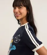 Remera azul marino de algodón con cuello redondo y mangas cortas con rayas blancas en los hombros. Estampado frontal de la Pitufina jugando al tenis.
