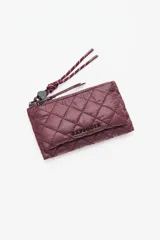 Billetera o monedero acolchado color vino tinto, con costuras en patrón de rombos. Presenta un cierre de cremallera y una correa de mano trenzada en tonos vino y blanco. El nombre de la marca está grabado en relieve en la parte frontal.