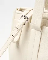 Cartera tipo tote de cuero sintético color crudo, con doble asa de mano, detalles de correas con hebilla y un charm colgante en forma de corazón. Cuenta con placa metálica con el logo de la marca en el frente.