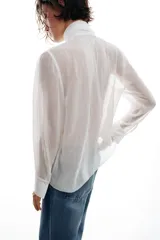 Blusa de tejido semitransparente con efecto arrugado, escote en pico y manga larga. Presenta un detalle decorativo frontal con estructura en forma de flor.