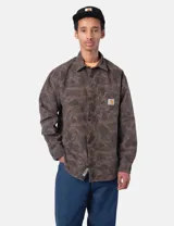 Camisa de manga larga Carhartt con estampado camuflado en tonos verdes y marrones. Presenta cuello clásico, cierre frontal con botones, un bolsillo en el pecho con etiqueta de la marca y puños abotonados.
