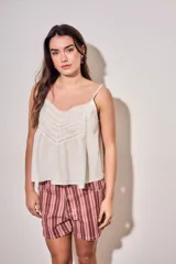 Blusa color off white de algodón peinado, sin mangas, con tirantes finos, escote en V con detalles de encaje y bordados, y pliegues decorativos en los laterales.
