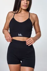 Conjunto deportivo de dos piezas en color negro, compuesto por un top con espalda descubierta y tiras cruzadas, y un short de tiro alto con efecto fruncido en la parte posterior. El short presenta el logo de la marca estampado en la cintura.