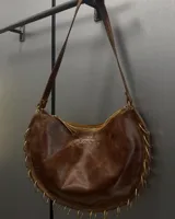 Cartera de cuero vacuno color marrón con diseño semicircular, detalle de argollas metálicas en el borde inferior y correa larga ajustable.