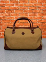 Bolso de viaje tipo duffel confeccionado en lona color tostado con detalles de cuero marrón en las esquinas, bordes superiores y asas. Presenta un diseño clásico con herrajes metálicos y un pequeño logo circular en el centro.