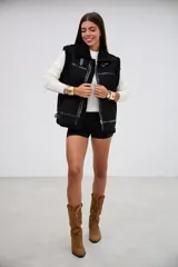 Chaleco negro de estilo aviador con interior y bordes de corderito sintético. Presenta cierre frontal metálico, detalles de tiras de cuero sintético y hebillas ajustables en el cuello y el ruedo.