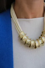 Collar de diseño artesanal compuesto por múltiples tiras de cuero en color crudo y piezas cilíndricas de metal dorado con textura martillada.