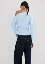 Blusa de algodón celeste con diseño asimétrico de un solo hombro, detalles fruncidos y manga larga.