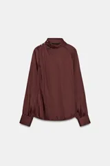 Blusa de tejido satinado color bordó, con cuello subido drapeado y manga larga con puños. Presenta detalle de pliegues frontales y cierre con botones laterales en el cuello.