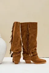 Botas altas estilo texana de gamuza color camel, con taco cuadrado y puntera fina. Presentan flecos largos colgando a lo largo del lateral de la caña y una correa con hebilla metálica y argollas alrededor del tobillo.