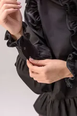 Camisa celeste con volados decorativos en los hombros, pecho, mangas y ruedo, destacados con ribetes en tono rojo. Presenta cierre frontal de botones, cuello alto y mangas largas abullonadas con puños ajustados.