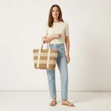 Bolso tipo tote confeccionado en paja tejida con diseño de rayas horizontales en tonos beige y marrón. Cuenta con doble asa de hombro de lona clara y cierre superior.