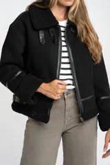 Campera tipo gamulán de corte oversize, color negro, con interior y detalles en cuello y puños de corderito sintético. Presenta cierre frontal y correas decorativas en el cuello.