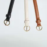Pack de tres cinturones finos de cuero sintético en colores negro, marrón y blanco, con hebilla dorada en forma de semicírculo.