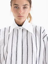 Camisa blanca de poplin con fit holgado, cuello con broche metálico y mangas largas con puños abotonados. Presenta un patrón de rayas verticales formadas por bordados de boquitas.