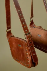 Cartera de mano tipo baguette confeccionada en cuero vacuno color marrón, con detalle de tachas y ojalillos metálicos en el frente. Cuenta con cierre superior, bolsillo trasero con cierre y forro interior de algodón.