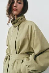 Cazadora de color verde militar, con cuello alto, manga larga abullonada con puños y cierre frontal con botones a presión ocultos. Incluye un cinturón ajustable del mismo tejido para entallar la cintura y bolsillos delanteros de vivo.