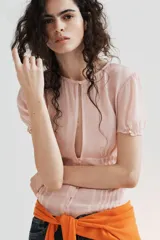 Top de georgette semitransparente color rosado, con cuello redondo, mangas cortas abullonadas y cierre frontal con botones. Presenta detalles de alforzas en la parte inferior.