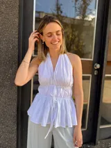 Blusa halter blanca sin mangas, con escote en V pronunciado, espalda descubierta, cintura fruncida con doble lazo frontal ajustable y diseño peplum con vuelo en la parte inferior.