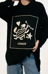 Sweater de punto oversize color crudo, con cuello redondo y diseño bordado a mano en el frente representando al signo Cáncer con un cangrejo, un corazón y una estrella.