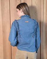 Camisa de denim celeste con cuello alto con volados, botonadura frontal y detalles de volados verticales a lo largo del frente. Cuenta con mangas largas abullonadas y puños con volados.