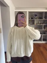 Buzo tejido de punto color tostado, con cuello redondo y diseño de trenzas verticales en el frente y mangas. Presenta un corte holgado con puños y ruedo acanalados.