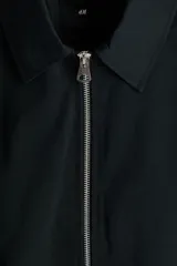 Campera negra de sarga de algodón, con cuello inglés, cierre frontal metálico y bolsillos delanteros ribeteados. Presenta elástico revestido en puños y bajo, y diseño forrado en su interior.