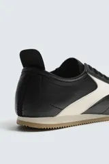 Zapatilla de estilo retro con cordones, confeccionada en una combinación de piezas de cuero sintético negro y detalles en blanco, con suela dentada de goma en tono caramelo.