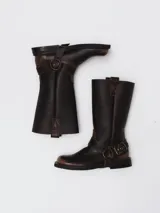 Bota de caña alta estilo western confeccionada en cuero con acabado avejentado. Presenta punta cuadrada, taco bajo y una correa con estribo metálico desmontable que rodea el tobillo.