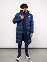 Parka larga acolchada color azul marino con capucha, cierre frontal con botones a presión y bolsillos laterales. Presenta el logo de Umbro y el escudo del Club Nacional de Football bordados en el pecho.