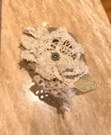 Prendedor artesanal con forma de flor, compuesto por capas de tejido de punto crochet color crema y tela de arpillera o tejido rústico color beige. En el centro tiene un dije metálico redondo con diseño grabado en tonos oscuros.