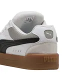 Championes Puma modelo Park Lifestyle SK8, de diseño urbano inspirado en el skate. Presentan una base de cuero blanco con detalles en gamuza gris claro en la puntera y el talón, destacando la icónica franja lateral de Puma en color negro. La suela es de goma color caramelo, proporcionando un contraste clásico y duradero.
