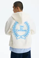 Campera con capucha H&M de felpa boxy en color beige, con cierre frontal, bolsillos tipo canguro y pequeño estampado azul en el pecho con la frase 'Not for Ceremony'.