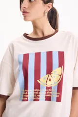 Remera de algodón blanca de manga corta, con corte suelto (loose fit) y rib en contraste marrón en el cuello y los puños. Presenta un estampado frontal rectangular con rayas verticales en tonos burdeos y celestes, superpuesto con el dibujo de una rodaja de limón amarilla y texto impreso.