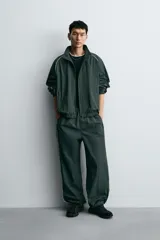 Pantalón jogger de corte relajado confeccionado en tejido técnico de color verde oscuro. Presenta cintura elástica con cordón interior, bolsillos laterales con cierre y bajo ajustable con elástico. Incluye detalles de líneas en contraste a lo largo de las piernas.