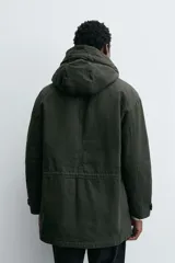Parka color verde militar de corte relaxed fit, confeccionada en tejido de algodón con interior acolchado. Cuenta con cuello subido, capucha ajustable con cordón, cierre frontal de cremallera oculta por solapa con botones a presión y amplios bolsillos frontales de plastrón con solapa.