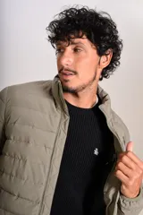 Campera puffer de corte clásico en color beige, con cierre frontal, cuello alto y puños elásticos. Presenta un diseño acolchado horizontal y un pequeño logo bordado en el pecho.