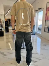 Remera color camel con cuello redondo y mangas cortas, con diseño gráfico estampado en la espalda que incluye un pájaro con sombrero de copa y la frase circular "Focus on finding joy in the journey".