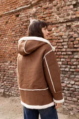 Campera estilo gamulán color tostado con interior y bordes de corderito sintético color crema. Presenta capucha amplia, cierre frontal con trabas y broches metálicos, y bolsillos delanteros grandes. Posee un calce relajado y diseño de textura suave.