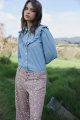 Camisa de denim azul claro con cuello clásico, cierre frontal de botones y detalle de volados en los hombros. Presenta mangas largas y un corte relajado.