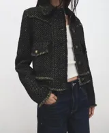 Chaqueta corta confeccionada en tejido texturizado con mezcla de algodón, poliéster y lana. Presenta cuello solapa, mangas largas, bolsillos frontales con solapa y cierre mediante botones metálicos dorados. Los bordes cuentan con detalles de flecos deshilachados.