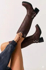 Bota de caña alta estilo biker, confeccionada en cuero negro. Presenta tres hebillas metálicas decorativas, cierre lateral y taco de madera.