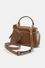 Bolso de mano tipo city color marrón, con diseño estructurado, costuras a la vista en color blanco y detalles de hebillas metálicas decorativas en la parte frontal. Cuenta con asa de mano superior, correa ajustable para usar como bandolera y cierre mediante solapa con imán.