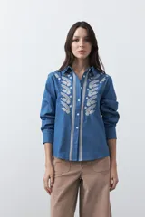 Camisa de manga larga en color azul oscuro con bordados florales en hilo blanco a lo largo de la botonadura frontal. Presenta cuello clásico y corte relajado.