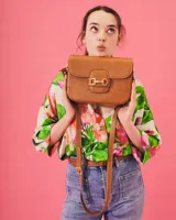 Cartera negra de cuero charolado con solapa y cierre de broche. Presenta un detalle metálico dorado en forma de freno de caballo en la parte frontal y una correa ajustable.