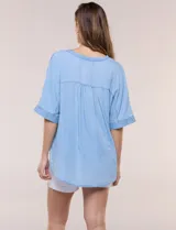 Blusa estilo denim de lyocell celeste, de silueta amplia y suelta, con escote en V, cartera frontal de botones y mangas cortas japonesas de hombros caídos. Cuenta con ruedo asimétrico y posibilidad de anudar al frente.