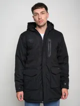 Parka larga negra con capucha y cierre frontal con cremallera. Tiene bolsillos laterales con solapa y un bolsillo vertical con cremallera en el pecho.